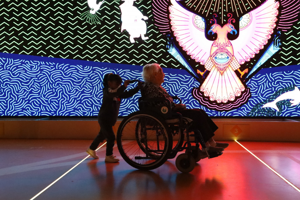 Niña de 5 años empuja la silla de ruedas de adulta mayor de 105 años de edad, al fondo se observa la pantalla luminosa de (leds) con un dibujo proyectado