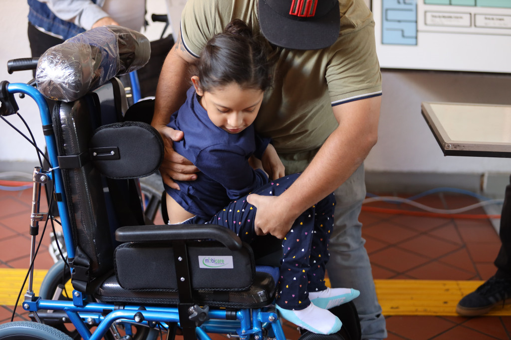 Padre de familia ayuda a su hija pequeña a colocarse en le silla de ruedas de parálisis cerebral infantil (PCI)