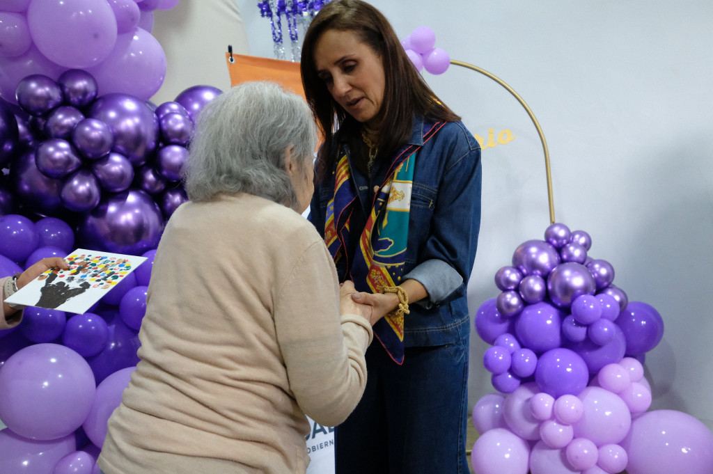 Sra. Maye conversa con una adulta mayor tomámdola de las masnos al fondo, la decoració con globos